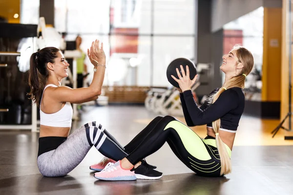 Dos jóvenes atléticas vestidas con ropa deportiva están haciendo juntos ejercicios deportivos para la prensa con pelota de fitness en el gimnasio moderno — Foto de Stock