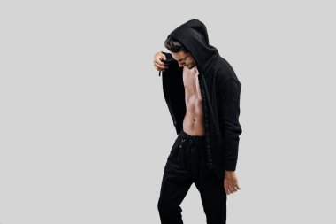 Şık genç dansçı bir kapüşonlu ve siyah pantolon ile çıplak bir gövde üzerinde bir sweatshirt giymiş beyaz bir arka plan üzerinde duruyor