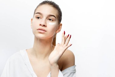 Beyaz giysiler giymiş makyaj olmadan güzel genç kahverengi saçlı kız beyaz arka plan üzerinde yüzüne bir krem koyar