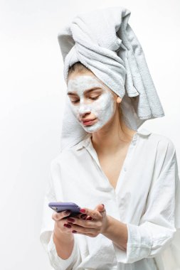 Saç ve kozmetik maskesi yüzünde beyaz bir havlu ile beyaz elbise giymiş güzel genç kadın ellerinin beyaz arka plan üzerinde cep telefonu tutar