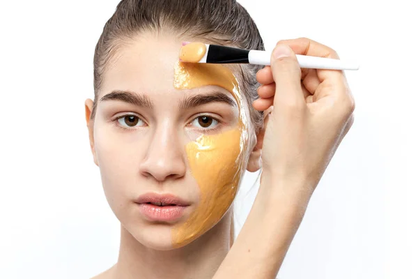 O processo de aplicação de máscara cosmética amarela com uma escova no rosto da jovem menina de cabelos castanhos — Fotografia de Stock
