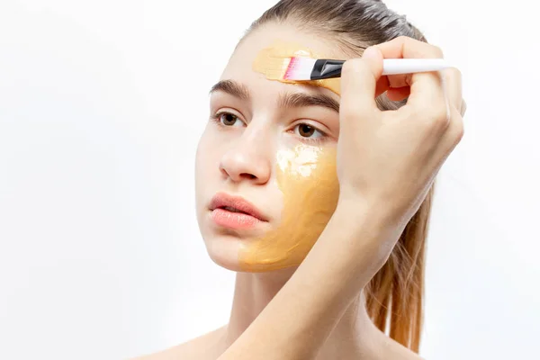 Prosessen med å påføre gul kosmetisk maske med en børste på ansiktet til ung, brunhåret jente – stockfoto