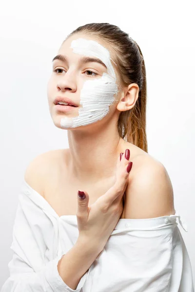 Young woman dressed in white clothes with a white cosmetic mask on a half of her face holds her hand at her shoulder on the white background — Stock Photo, Image