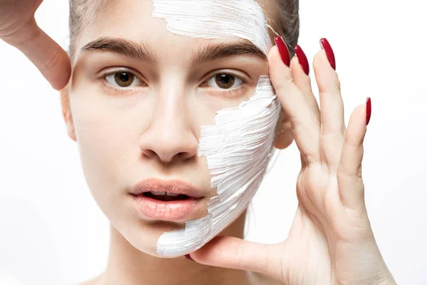 Egy fiatal nő, fehér kozmetikai maszkkal az arcán tartja a kezét az arcához a fehér háttérben. — Stock Fotó