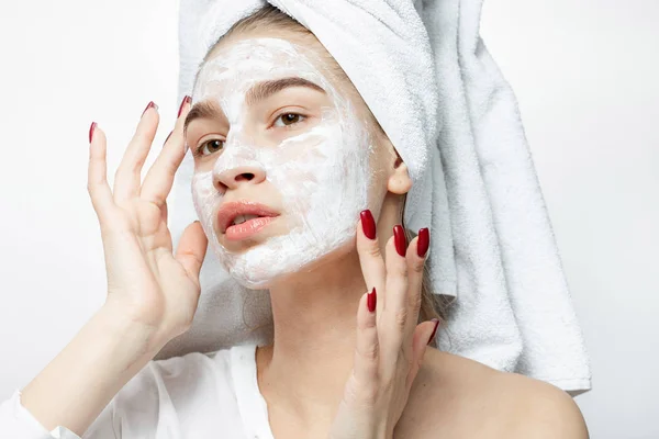 Menina agradável em uma roupa branca com uma toalha branca em seu cabelo coloca uma máscara cosmética em seu rosto — Fotografia de Stock