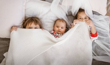 İki küçük kız kardeş ve küçük bir erkek kardeşi hafif rahat yatak odasında beyaz battaniye altında yatakta yalan