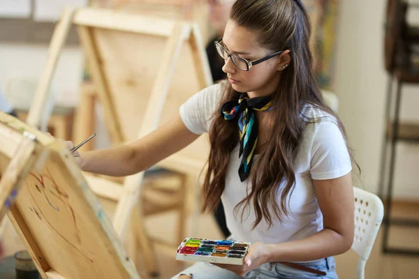 Menina de cabelos castanhos jovem em óculos vestidos com t-shirt branca e jeans com um lenço em torno de seu pescoço pinta um quadro no estúdio de arte — Fotografia de Stock