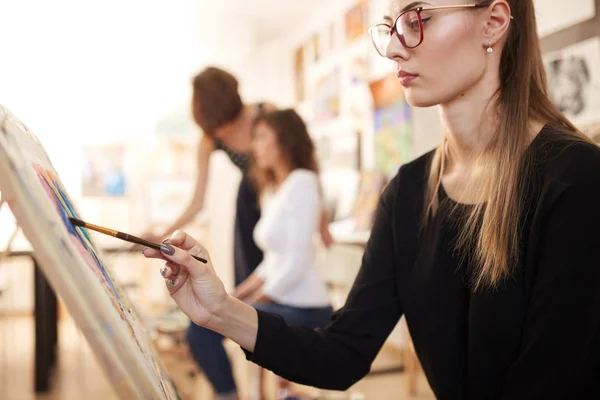 Pěkná mladá dívka v brýlích, oblečená v černé košile a džíny sedí na stojan a maluje obraz ve škole kreslení — Stock fotografie