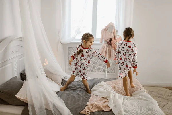 Дві маленькі дівчата в піжамі весело стрибають на ліжку в сонячній затишній спальні — стокове фото