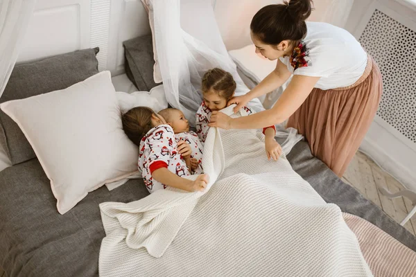 Młoda matka przykrywa biały koc dla dwóch córek i małego synka, który leży na łóżku w jasnej, przytulnej sypialni — Zdjęcie stockowe