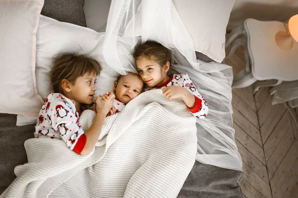 Δύο μικρές αδελφές και ένας μικροσκοπικός αδελφός ξαπλώσετε στο κρεβάτι κάτω από τη λευκή κουβέρτα στο φωτεινό άνετο υπνοδωμάτιο — Φωτογραφία Αρχείου