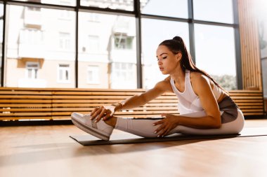 Muhteşem ince koyu saçlı kız beyaz spor üst ve tayt giyinmiş spor salonunda fitness için mat germe yapıyor