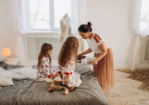 Молода красива мати приносить какао з зефіром та печивом дочкам, які сидять на ліжку в повному світлому затишному номері — стокове фото