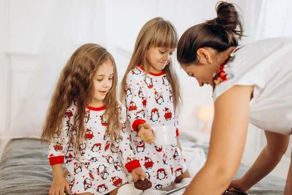 Młoda matka przynosi kakao z piankami i ciasteczkami córkom w piżamie, siedzącym na złym w jasnym, przytulnym pokoju. — Zdjęcie stockowe