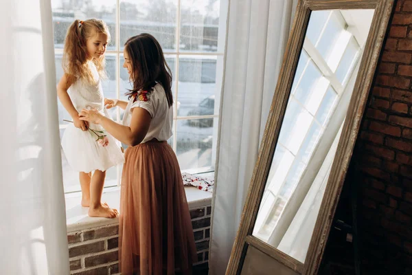 Молодая мать держит свою маленькую дочь стоя на подоконнике рядом с зеркалом в полной легкой уютной комнате — стоковое фото