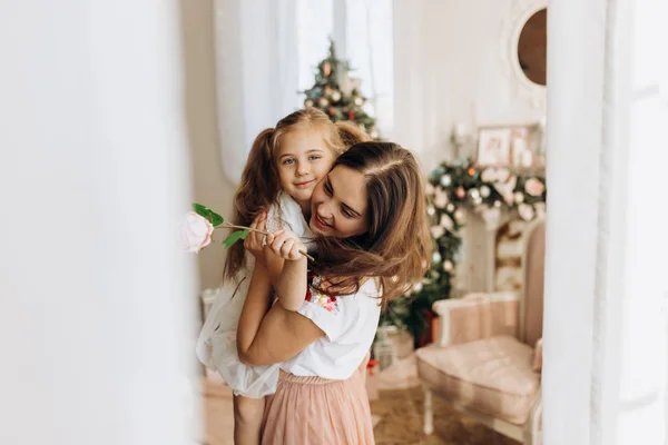 Молода мама тримає на руках свою маленьку доньку в затишній кімнаті з новорічною ялинкою — стокове фото
