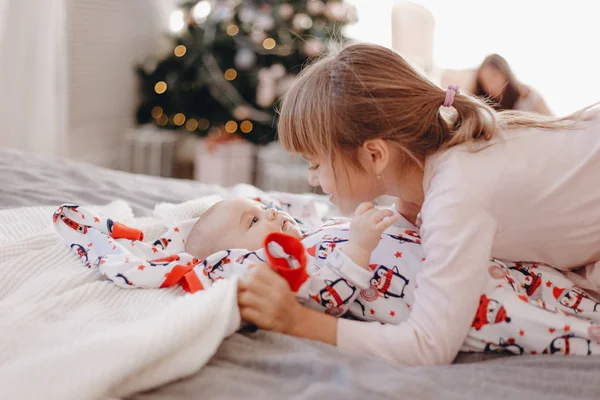 Маленька дівчинка, одягнена в піжаму, дивиться на свого маленького брата, який лежить на ліжку в затишній кімнаті з новорічною ялинкою — стокове фото