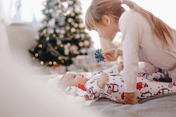 Dziewczynka ubrana w piżamę patrzy na jej maleńkiego brata leżącego na łóżku w przytulnym pokoju z drzewem nowego roku — Zdjęcie stockowe