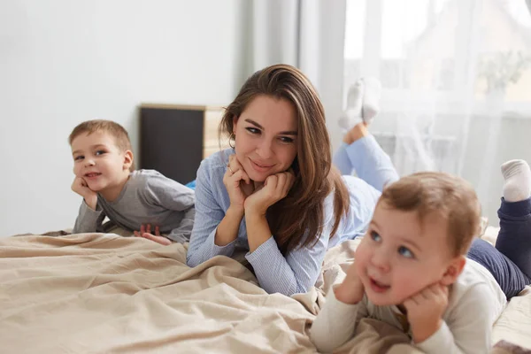 Szczęśliwa młoda matka, ubrana w piżamy niebieskie światło kładzie z jej dwóch synów mały, trzymając ręce pod ich podbródki na łóżku z beżowy kocyk w sypialni z dużym oknem — Zdjęcie stockowe