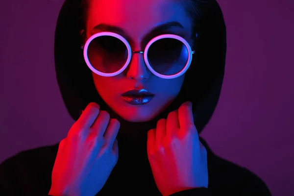 Retrato de niña de moda en un suéter negro con capucha y gafas de sol redondas en luz de neón roja y azul en el estudio —  Fotos de Stock