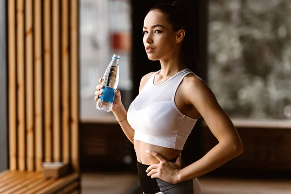 흰색 스포츠 탑을 입고 아름다운 슬림 검은 머리 소녀는 체육관에서 물 병을 보유 — 스톡 사진