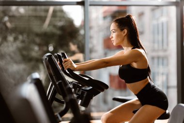 Kahverengi saçlı, siyah spor bluzlu ve şortlu genç atletik kız spor salonunda egzersiz bisikletinde çalışıyor.