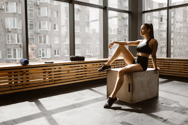 Encantadora chica calumnia vestida con top deportivo negro y pantalones cortos está sentado en una caja de madera a la luz del sol en frente de la ventana en el gimnasio — Foto de Stock