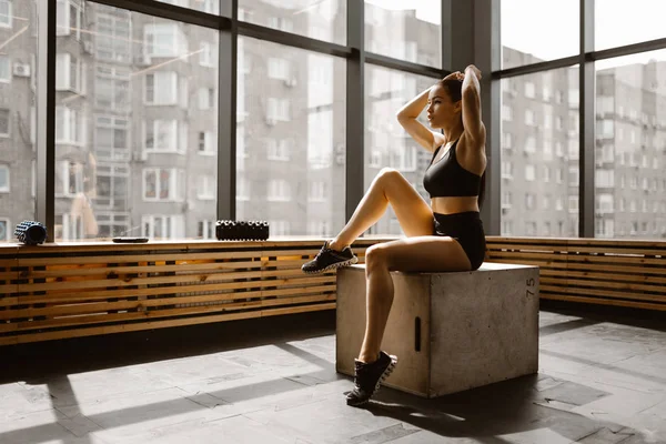 Hermosa chica delgada vestida con top deportivo negro y pantalones cortos está sentada en una caja de madera a la luz del sol en frente de la ventana en el gimnasio — Foto de Stock