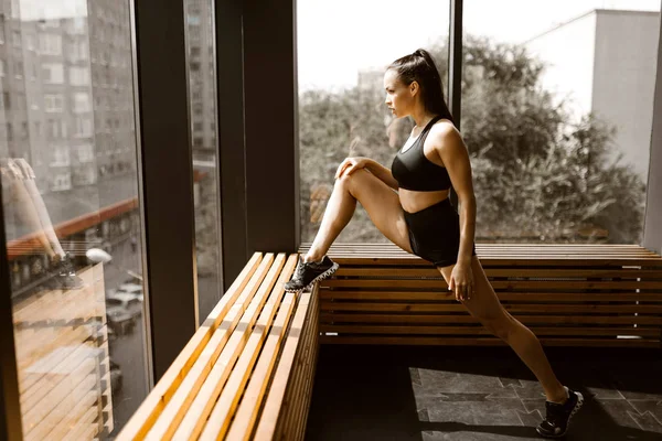 Hermosa chica calumnia vestida con top deportivo negro y pantalones cortos está haciendo estiramiento en un alféizar de ventana de madera en el gimnasio — Foto de Stock