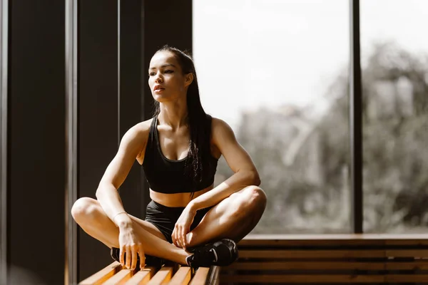Slanke brunette meisje gekleed in zwarte sporttop en shorts zit in Lotus pose op een houten vensterbank in de sportschool — Stockfoto