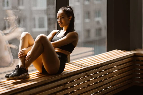 Atletische donkerharige meisje gekleed in zwarte sporttop en shorts zit op een houten vensterbank in de sportschool — Stockfoto