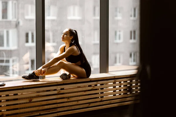 Atlético morena chica vestida con negro deportes top y pantalones cortos está sentado en un alféizar de madera ventana en el gimnasio — Foto de Stock