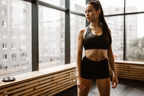 Hermosa chica atlética con el pelo castaño vestido con top deportivo negro y pantalones cortos se encuentra en el gimnasio — Foto de Stock