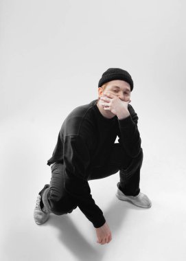 Siyah kot pantolon, kazak, şapka ve gri spor ayakkabı giymiş Adam çömelme ve beyaz arka plan üzerinde stüdyoda elleriyle yüzünü kapatıyor