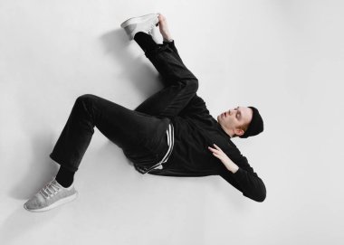 Freestyle dancer dressed in black jeans, sweatshirt, hat and gray sneakers is dancing lying on the floor in the studio on the white background clipart