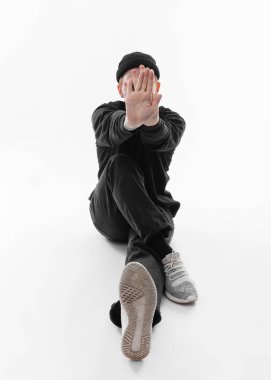 Siyah kot pantolon, sweatshirt, şapka ve gri spor ayakkabı giymiş Serbest stil dansçı beyaz arka plan üzerinde stüdyoda katta oturan dans ediyor