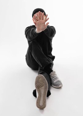Siyah kot pantolon, sweatshirt, şapka ve gri spor ayakkabı giymiş Serbest stil dansçı beyaz arka plan üzerinde stüdyoda katta oturan dans ediyor