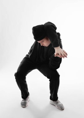 Siyah kot pantolon, sweatshirt, şapka ve gri spor ayakkabı giymiş Serbest stil dansçı beyaz arka plan üzerinde stüdyoda çömelme dans ediyor