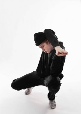 Siyah kot pantolon, sweatshirt, şapka ve gri spor ayakkabı giymiş Serbest stil dansçı beyaz arka plan üzerinde stüdyoda çömelme dans ediyor