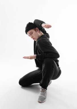Siyah kot pantolon, sweatshirt, şapka ve gri spor ayakkabı giymiş Serbest stil dansçı beyaz arka plan üzerinde stüdyoda çömelme dans ediyor