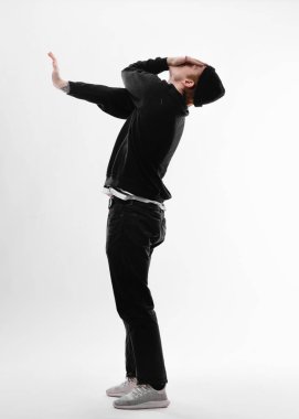 Siyah kot pantolon, sweatshirt, şapka ve gri spor ayakkabılar giymiş serbest dans eden genç dansçı, elini stüdyoda beyaz arka planda kapayarak dans ediyor.