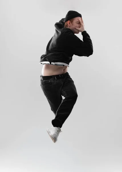 Cara vestida com jeans preto, capuz, chapéu e tênis cinza está pulando no estúdio no fundo branco — Fotografia de Stock