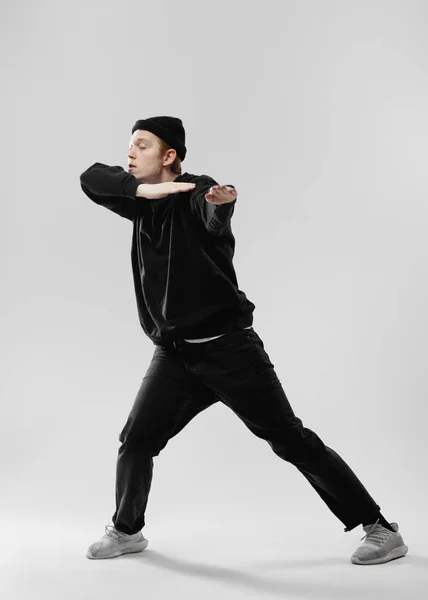 Tanečnice v černých džínách, mikina, čepice a šedé tenisky tancuje dělat pohyby rukama v ateliéru na bílém pozadí — Stock fotografie