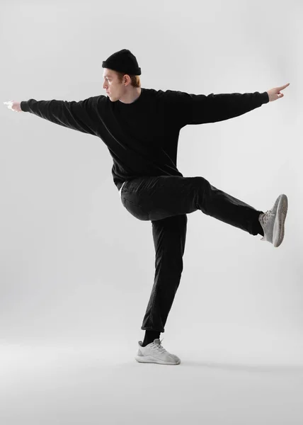 Dançarino vestido de jeans preto, camisola, chapéu e tênis cinza está dançando fazendo movimentos com as pernas no estúdio no fundo branco — Fotografia de Stock