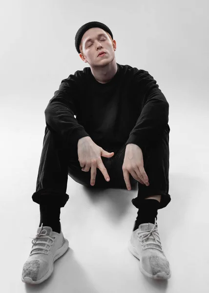 검은 청바지, 스웨트 셔츠, 모자와 회색 운동화를 입은 남자가 흰색 배경의 스튜디오 바닥에 앉아 있습니다. — 스톡 사진