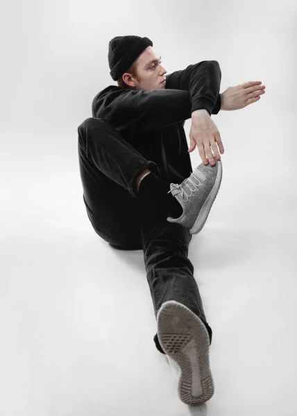 Freestyle-Tänzerin in schwarzen Jeans, Sweatshirt, Hut und grauen Turnschuhen tanzt sitzend auf dem Boden im Studio auf weißem Hintergrund — Stockfoto