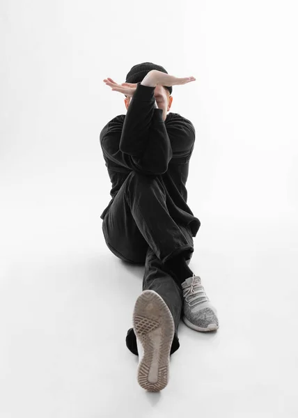 Akrobatický tanečník oblečený v černých džínách, tričku, klobouku a šedých teniskách je tanec sedící na podlaze ve studiu na bílém pozadí — Stock fotografie