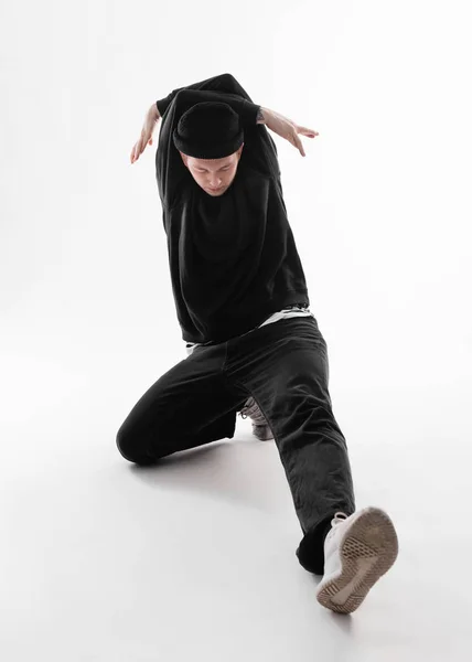 Freestyle dançarina vestida com jeans pretos, camisola, chapéu e tênis cinza está dançando agachamento no estúdio no fundo branco — Fotografia de Stock