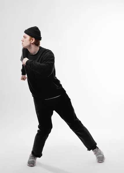 Jonge freestyle danser, gekleed in zwarte jeans, trui, hoed en grijze sneakers is dansen in de studio op de witte achtergrond — Stockfoto