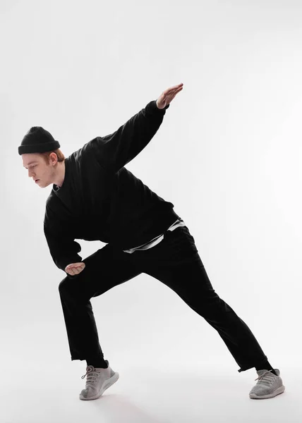 Молодий танцюрист, одягнений в чорні джинси, светр, капелюх і сірі кросівки, танцює вільний стиль в студії на білому тлі — стокове фото